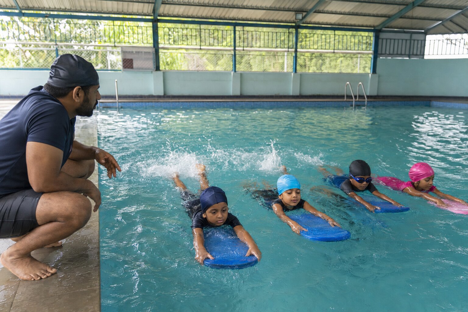Illustrative scene of children practising kickboard skills under a swimming coach's supervision